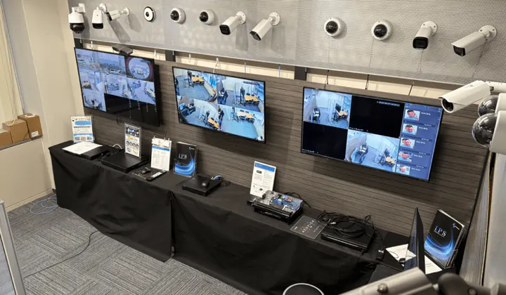 福岡オフィスのショールームの展示の壁。上段にカメラが並び、3台のモニターに映像、黒い布を掛けた長机に録画機とカタログ